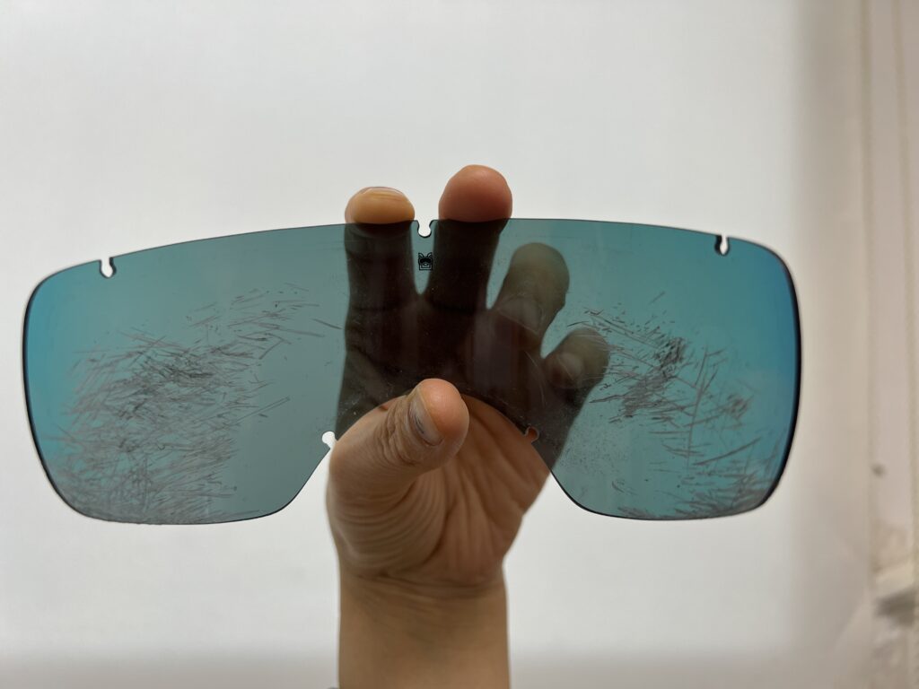 A scratched Kyunglas dog goggle lens being held up close, showing visible surface damage from pet use